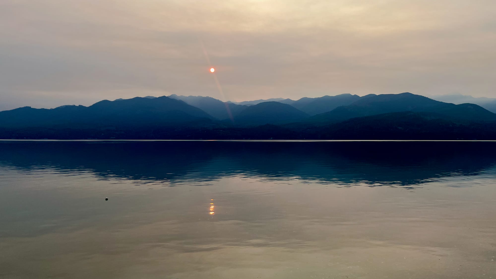 A distant sunset in a dusty, cloudy sky above the mountains and beyond a body of water.