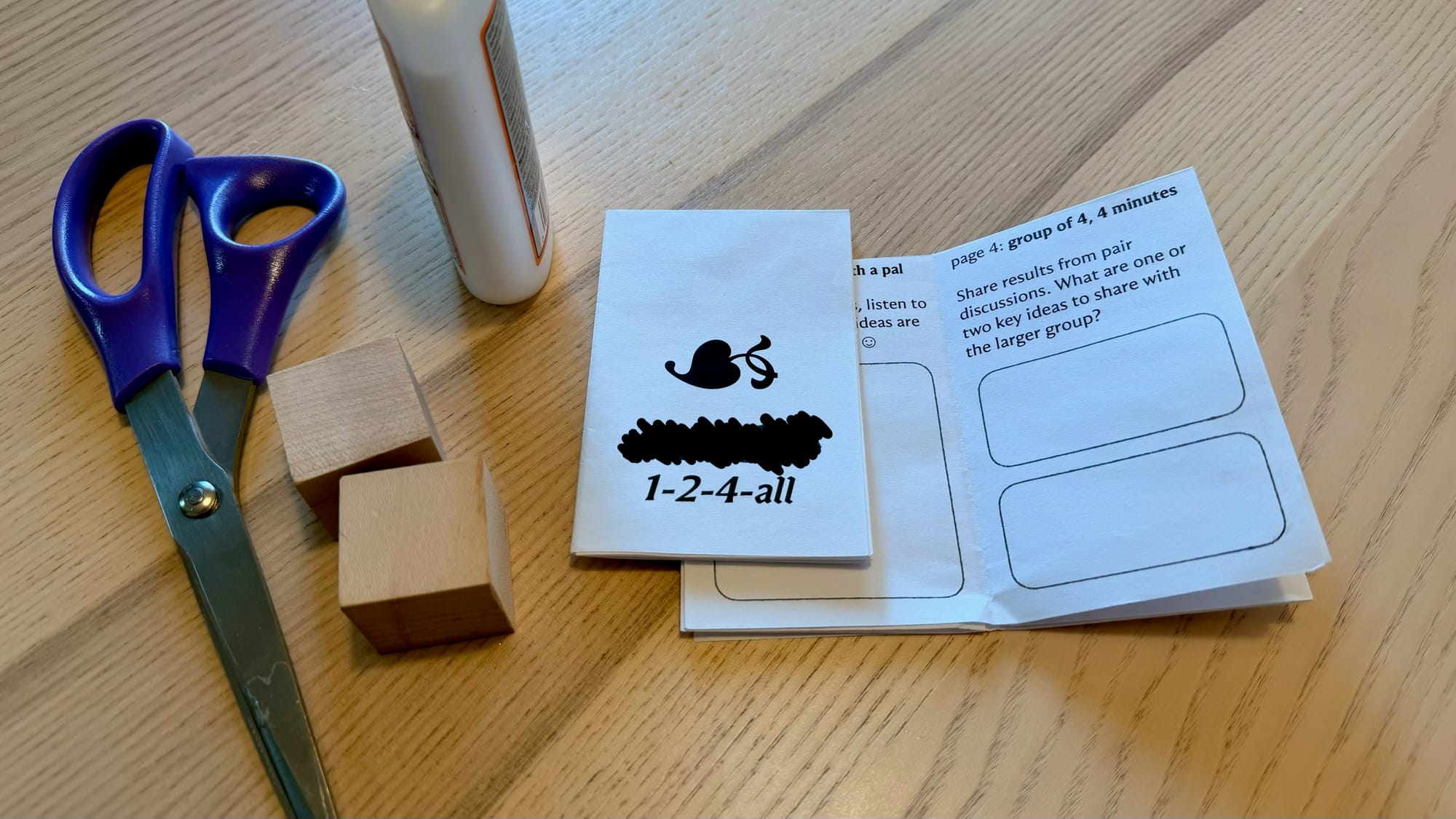 Two printed and folded mini-zines on a tabletop next to a bottle of glue, two wooden blocks, and a pair of scissors.
